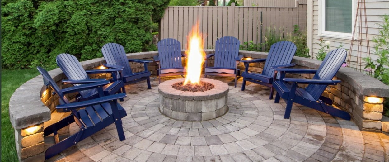A paver patio with a built-in fire pit and seating wall, lit for evenings