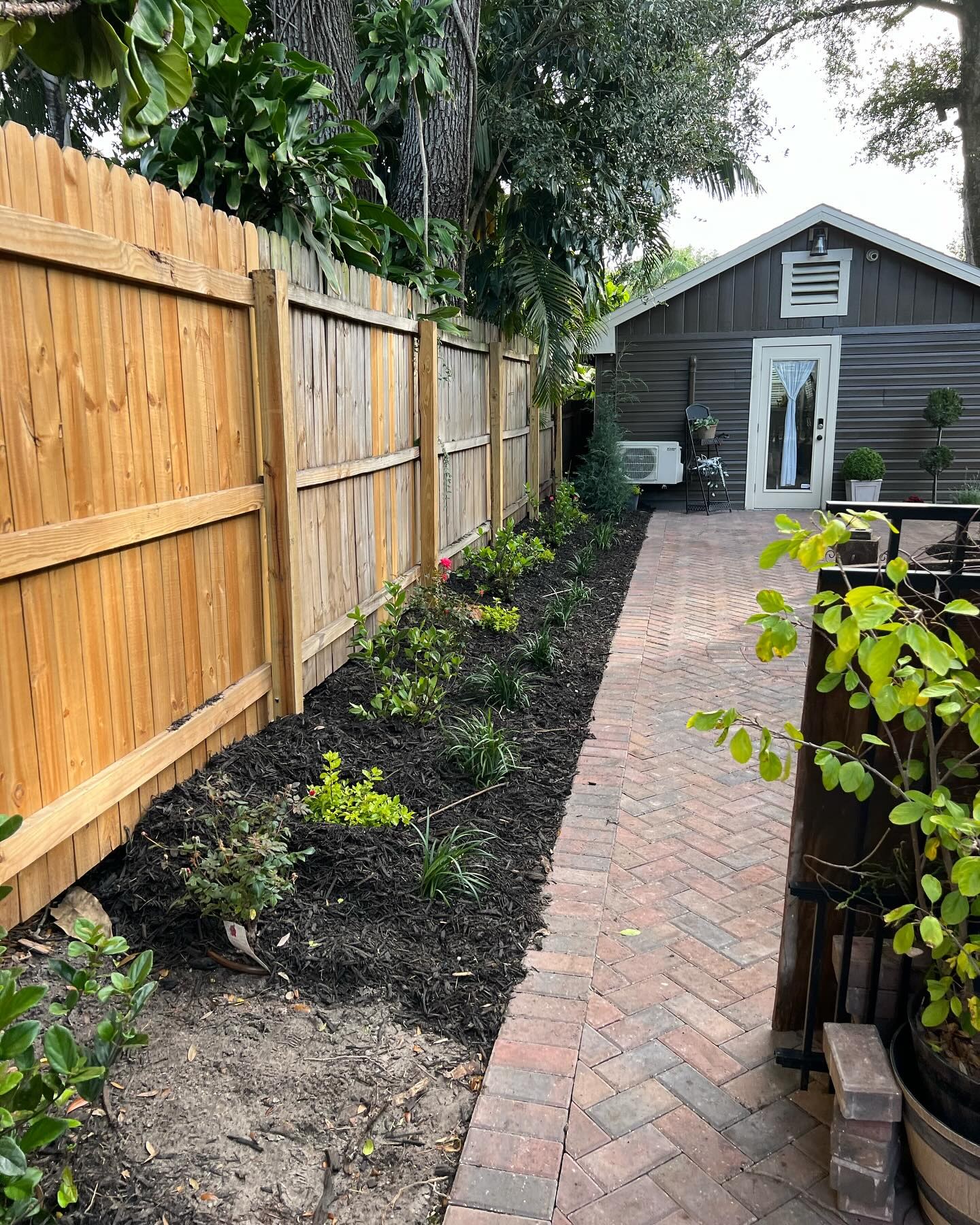 A brick paver path along a wood privacy fence, with new plantings softening the fence line