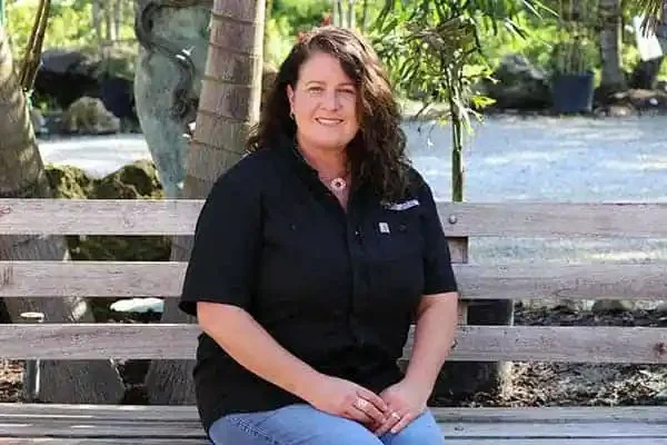 Tamara Slayton, President of Millennium Lawn, Landscape & Nursery, smiling on a wooden bench outdoors with palm trees and greenery behind her
